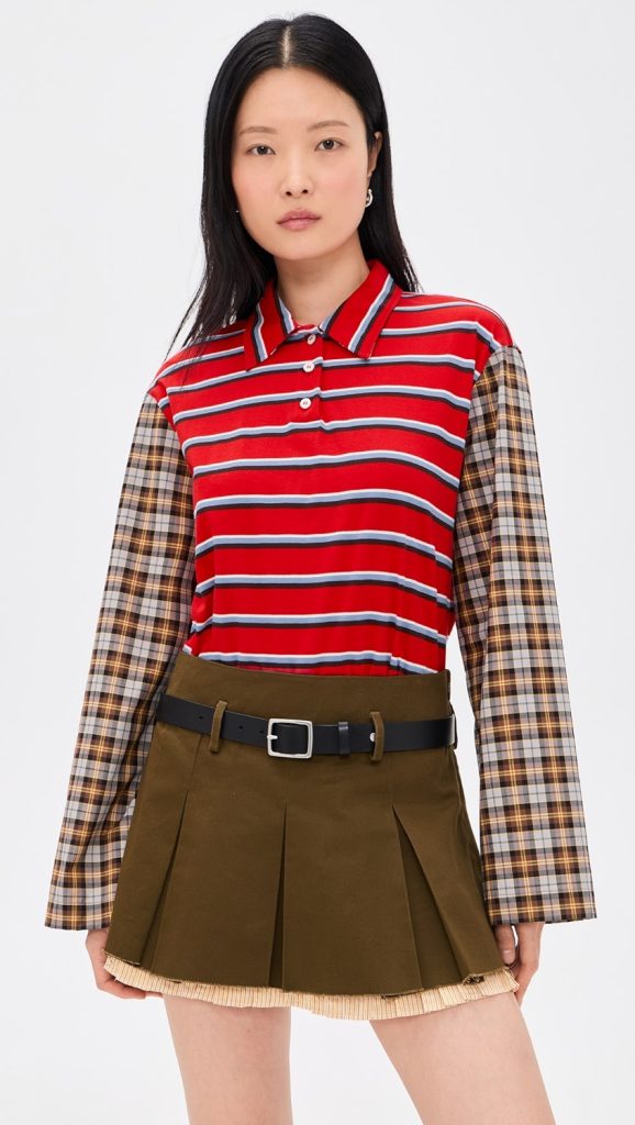 Woman wearing a red striped polo shirt with plaid sleeves and a brown pleated mini skirt with a black belt.