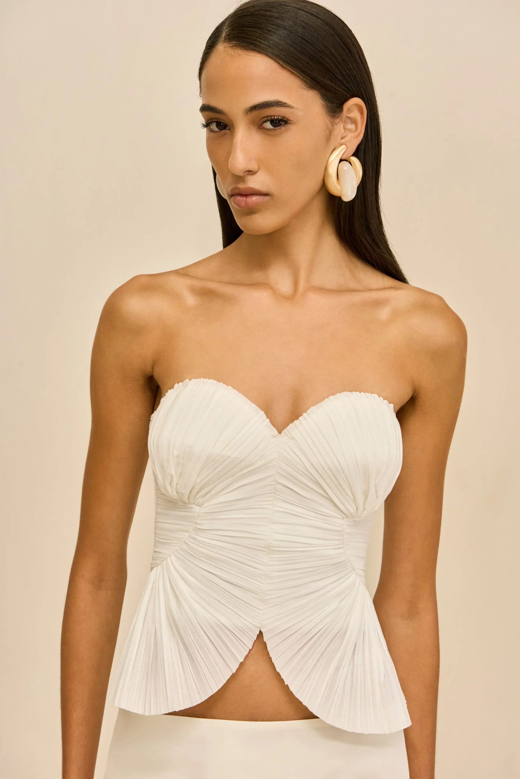 IMG_1379 - New Trend Model wearing a strapless white pleated gown with oversized cream earrings, facing slightly left against a beige backdrop.