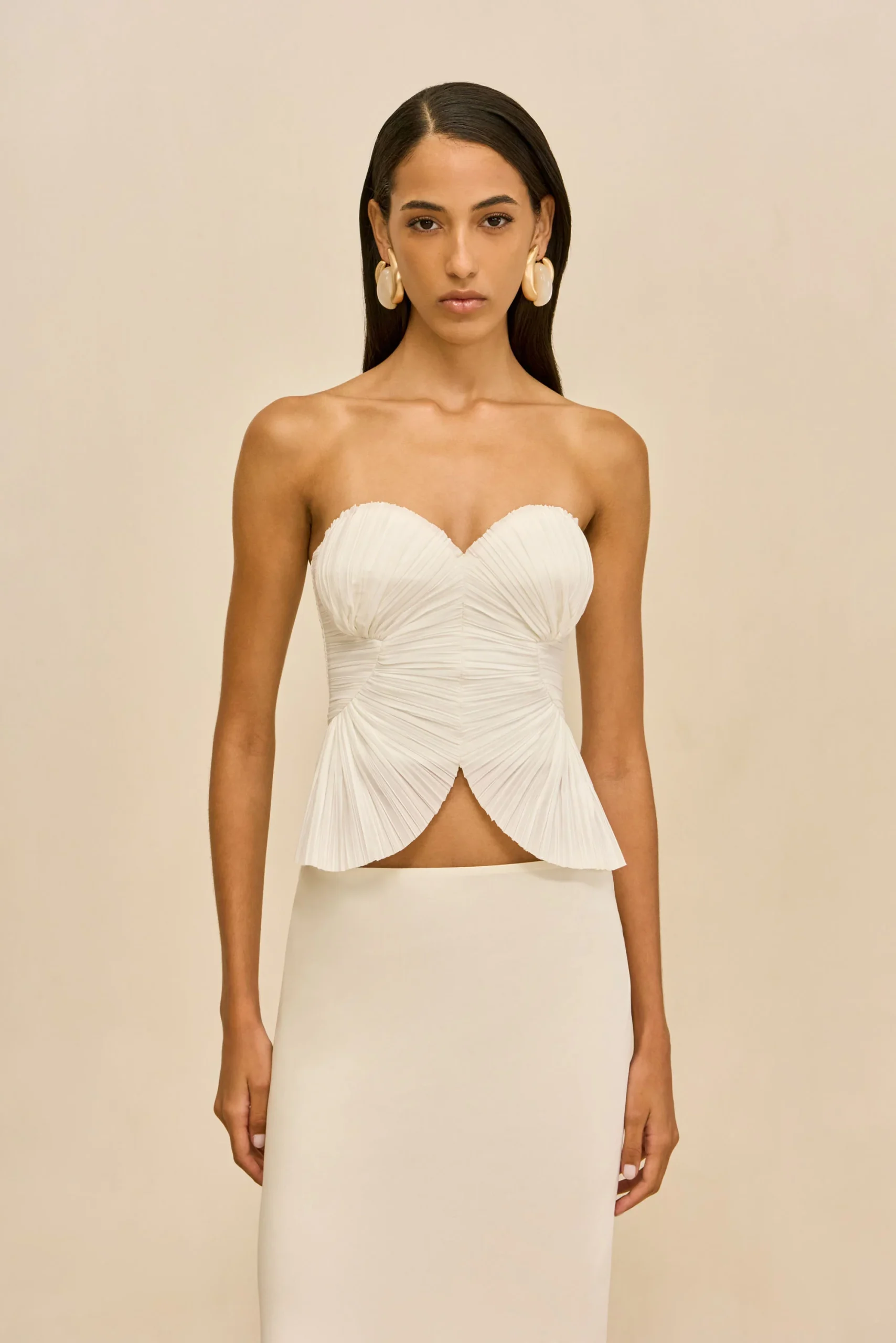 IMG_1380 - New Trend Model wearing a white strapless gown with a ruched, pleated bodice and a high-low skirt against a beige backdrop, accessorized with large cream earrings.