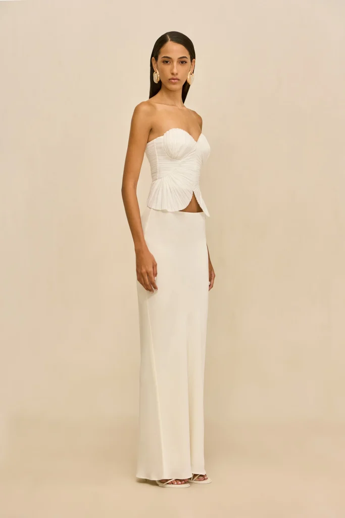 Model in a white strapless gown with a peplum waist and long skirt, standing against a beige backdrop, wearing large cream earrings.
