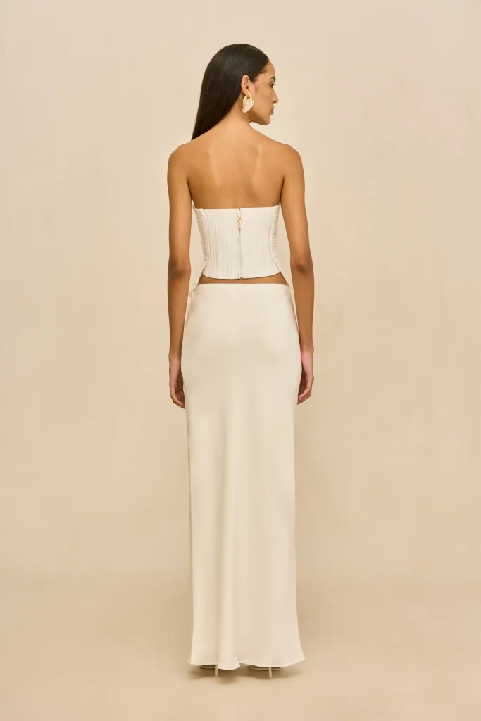 Back view of a woman in a white strapless gown with a corseted back and a long, flowing skirt, standing against a beige background.