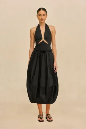 Female model wearing a black halter-neck dress with front cutouts and a voluminous midi skirt, paired with black sandals.