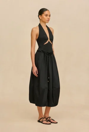 Model wearing a black halter-neck dress with cutouts at the waist and a gathered midi skirt, standing against a beige backdrop.