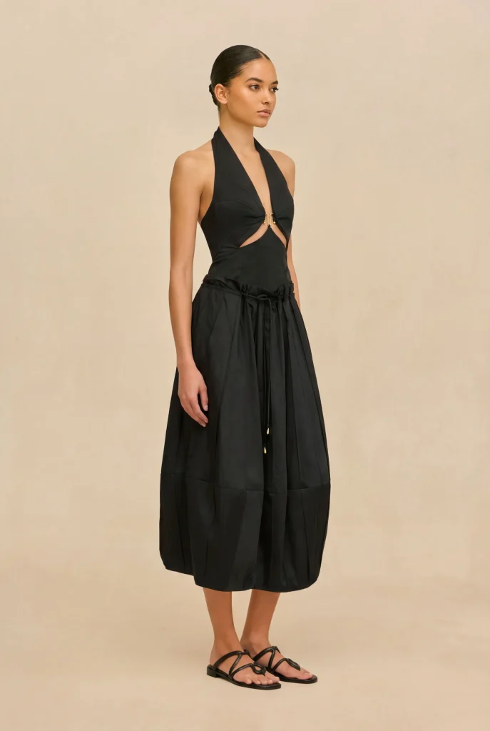 Model wearing a black halter-neck dress with cutouts at the waist and a gathered midi skirt, standing against a beige backdrop.
