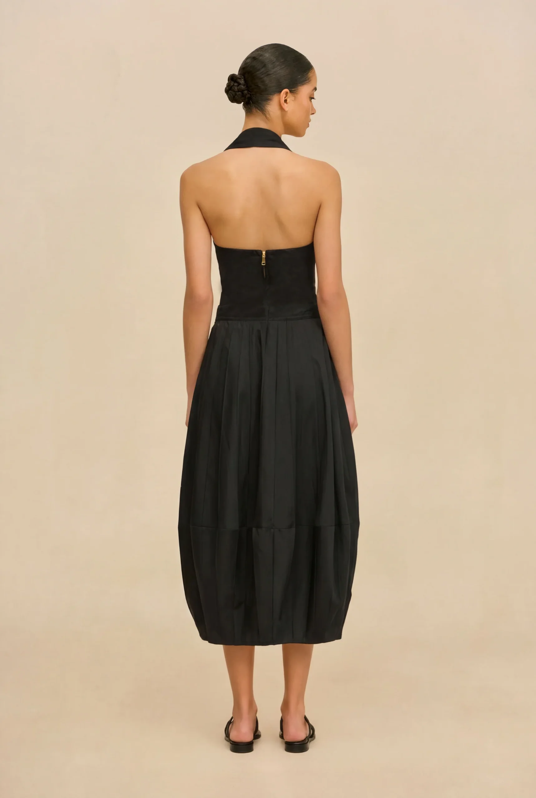IMG_1388 - New Trend Back view of a woman in a black halter-neck dress with a center-back zipper, against a beige backdrop.