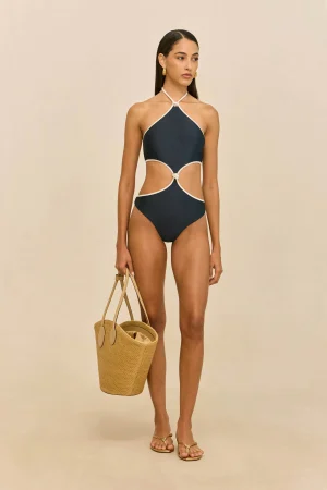 Model wearing a navy cut-out halter swimsuit with white trim, holding a straw tote bag against a beige backdrop.