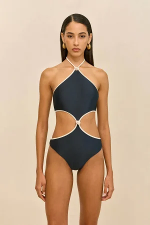 Model wearing a navy halter-neck one-piece swimsuit with white trim, standing against a beige background.