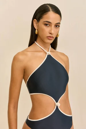 Portrait of a woman wearing a navy halter-neck swimsuit with white trim and gold earrings, against a beige background.