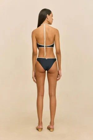 Woman shown from the back wearing a navy bikini with white trim, halter top tied at the neck, gold hoop earrings, standing against a beige background.