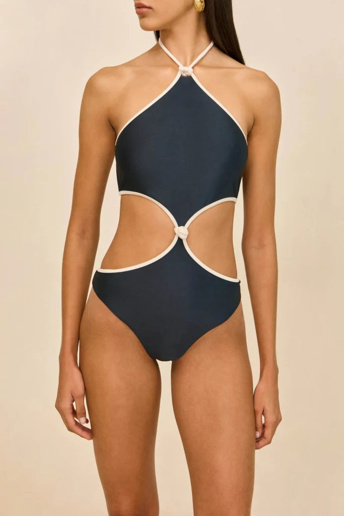 Woman wearing a navy blue one-piece swimsuit with white trim and knot accents at the neck and waist, standing against a beige background.