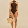 IMG_1395 - New Trend Model wearing a dark brown one-piece swimsuit with a twisted halter neckline, standing against a beige backdrop, carrying a woven straw bag.