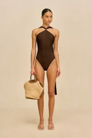 Model wearing a dark brown one-piece swimsuit with a twisted halter neckline, standing against a beige backdrop, carrying a woven straw bag.