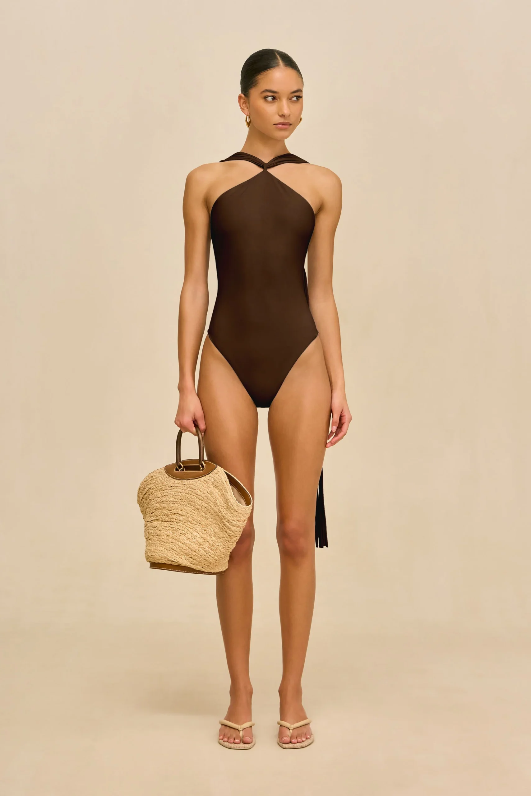 IMG_1395 - New Trend Model wearing a dark brown one-piece swimsuit with a twisted halter neckline, standing against a beige backdrop, carrying a woven straw bag.