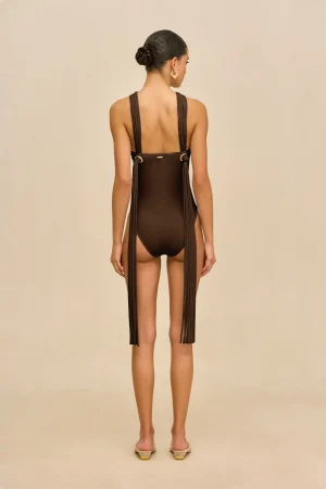 Back view of a woman wearing a brown one-piece swimsuit with long fringe panels down the sides, her hair in a neat bun, and gold hoop earrings, standing against a beige background.