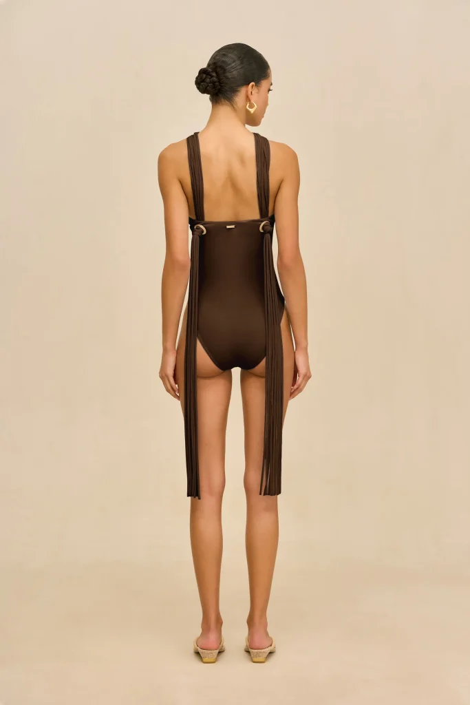 Back view of a woman wearing a brown one-piece swimsuit with long fringe panels down the sides, her hair in a neat bun, and gold hoop earrings, standing against a beige background.
