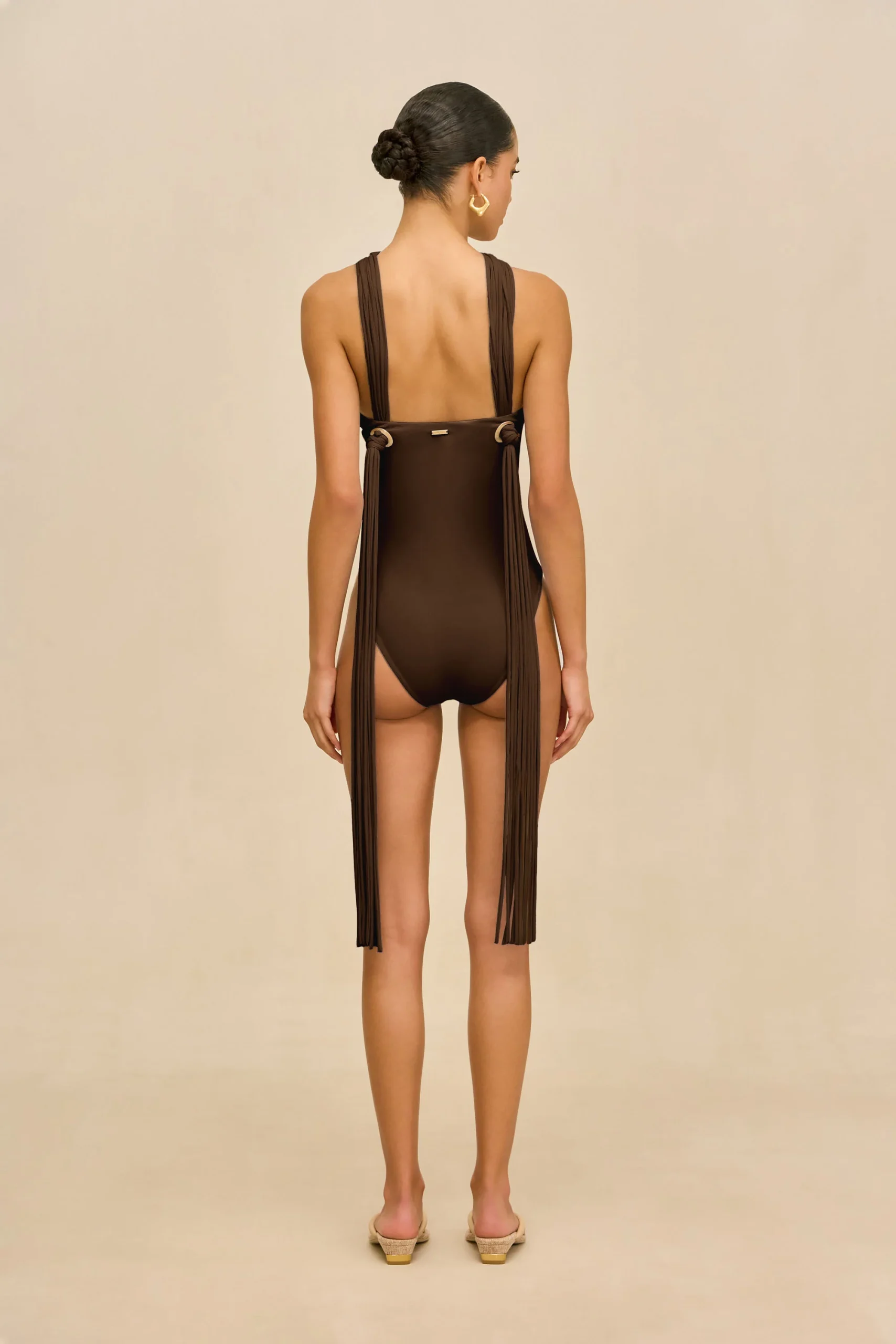 IMG_1396 - New Trend Back view of a woman wearing a brown one-piece swimsuit with long fringe panels down the sides, her hair in a neat bun, and gold hoop earrings, standing against a beige background.