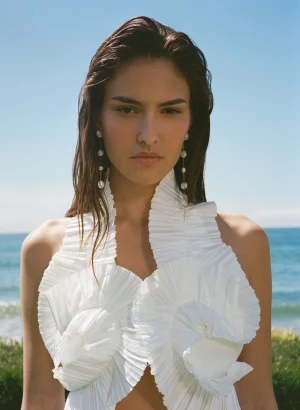 Woman in a white pleated, ruffled dress at the beach, looking at the camera with a calm expression; sea and blue sky in the background.