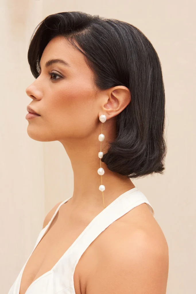 Profile view of a woman with dark hair wearing long pearl drop earrings and a white sleeveless top against a beige background.