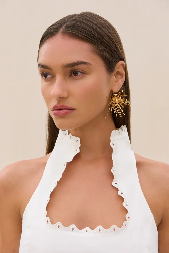 Woman with straight brown hair wearing a large gold sunburst earring and a white scalloped halter top against a beige background.