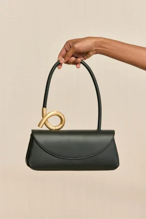 Dark gray handbag with a gold circular handle being held by a hand against a beige background.
