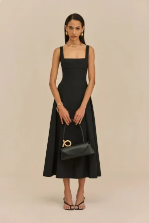 Full-length portrait of a woman in a black sleeveless square-neck dress with gold jewelry, holding a black handbag, standing barefooted? wearing sandals against a beige backdrop.