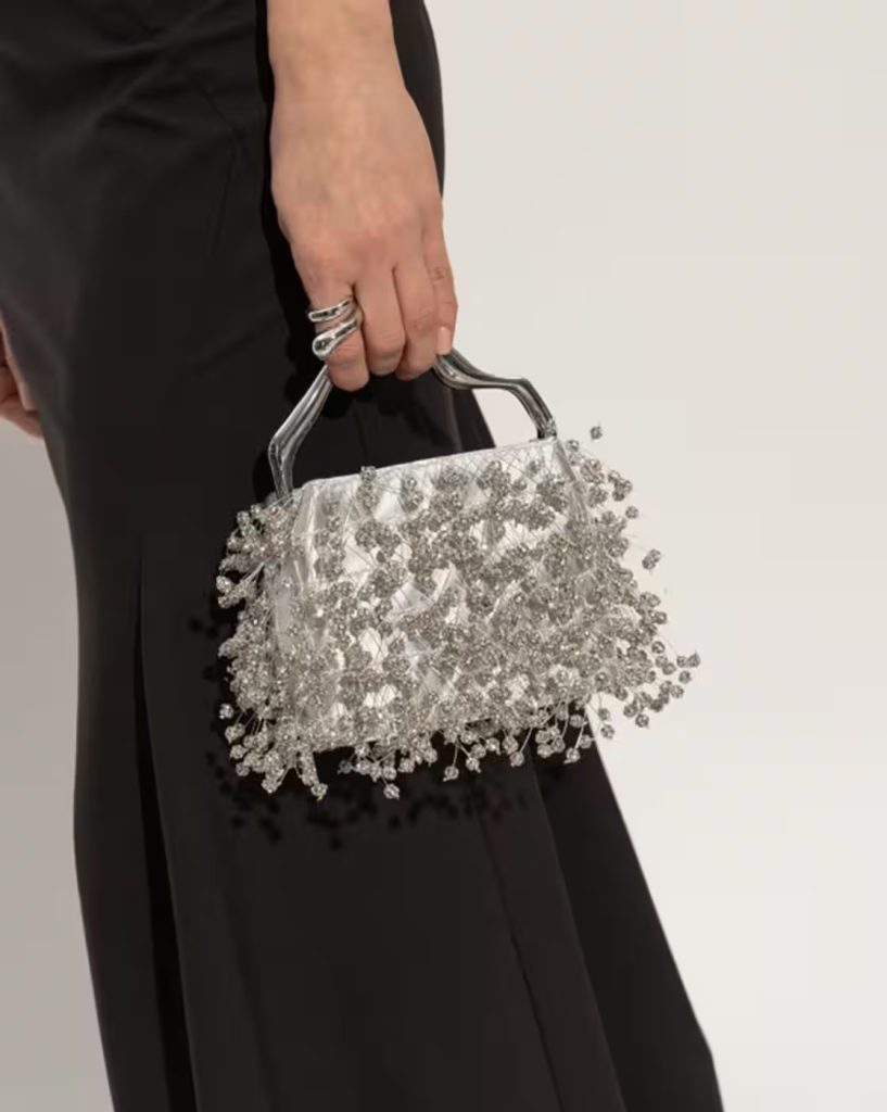A hand holding a silver beaded clutch with dangling crystal beads against a black dress.