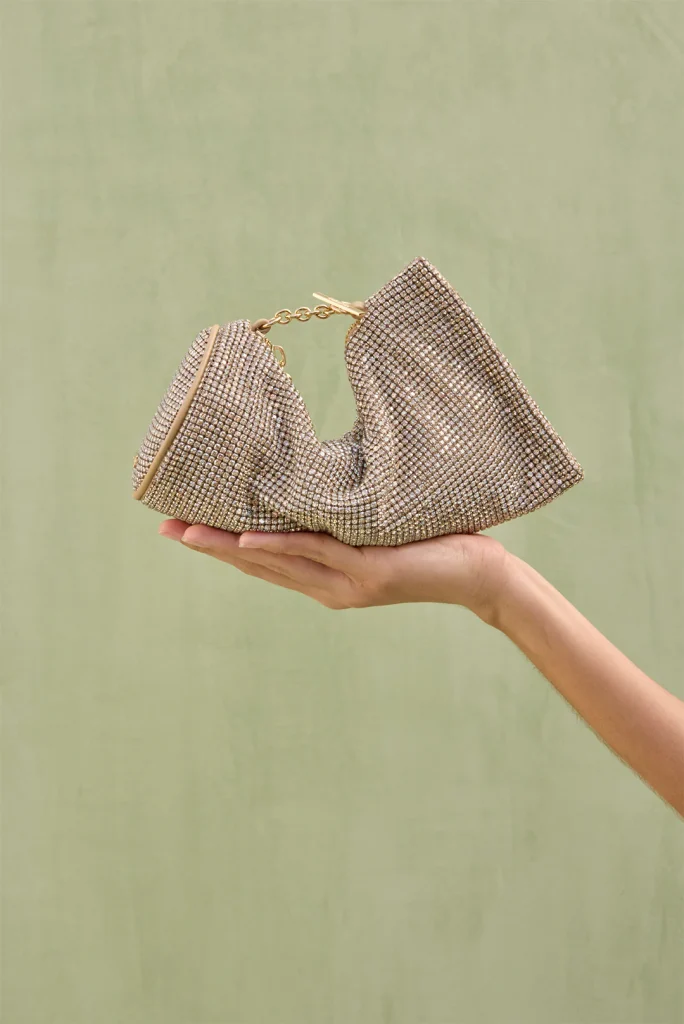 Sparkling gold-toned rhinestone clutch bag held in a palm against a pale green background with a short chain strap visible.