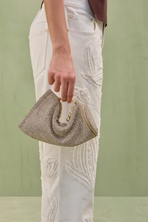 Hand holds a gold-toned beaded clutch against cream embroidered jeans in a pale outdoor setting.