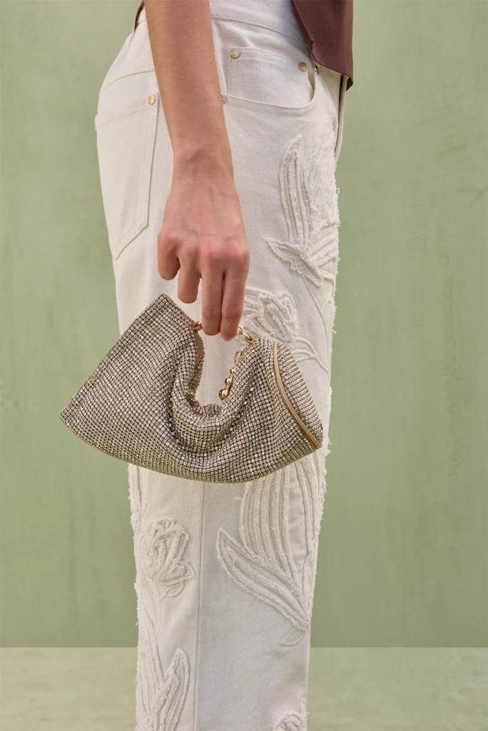 Hand holds a gold-toned beaded clutch against cream embroidered jeans in a pale outdoor setting.