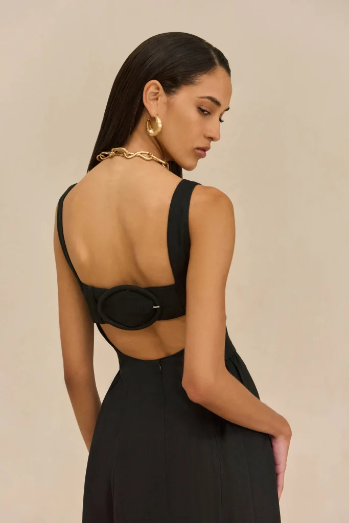 Side view of a woman in a black open-back dress wearing a chunky gold chain necklace and gold hoop earring against a beige background.