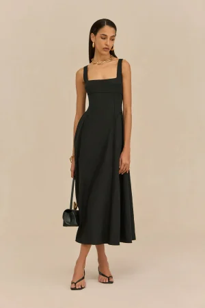 Woman wearing a black, square-neck dress with wide straps, gold jewelry, and strappy heels, standing against a neutral background.