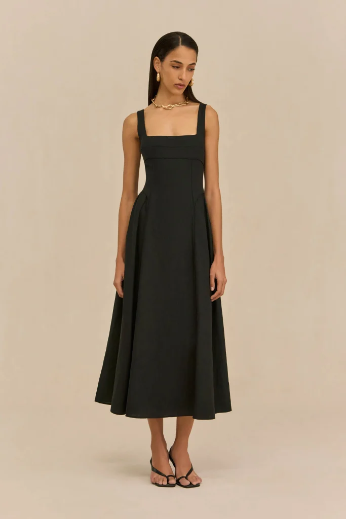 Woman in a black square-neck dress with gold jewelry, standing against a beige backdrop.