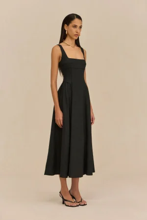Full-length image of a model in a black sleeveless midi dress with wide straps, gold necklace and hoop earrings, standing against a beige backdrop.