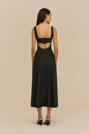Back view of a woman in a black sleeveless dress with an open lower back and a large oval belt, wearing a chunky gold necklace and hoop earrings.