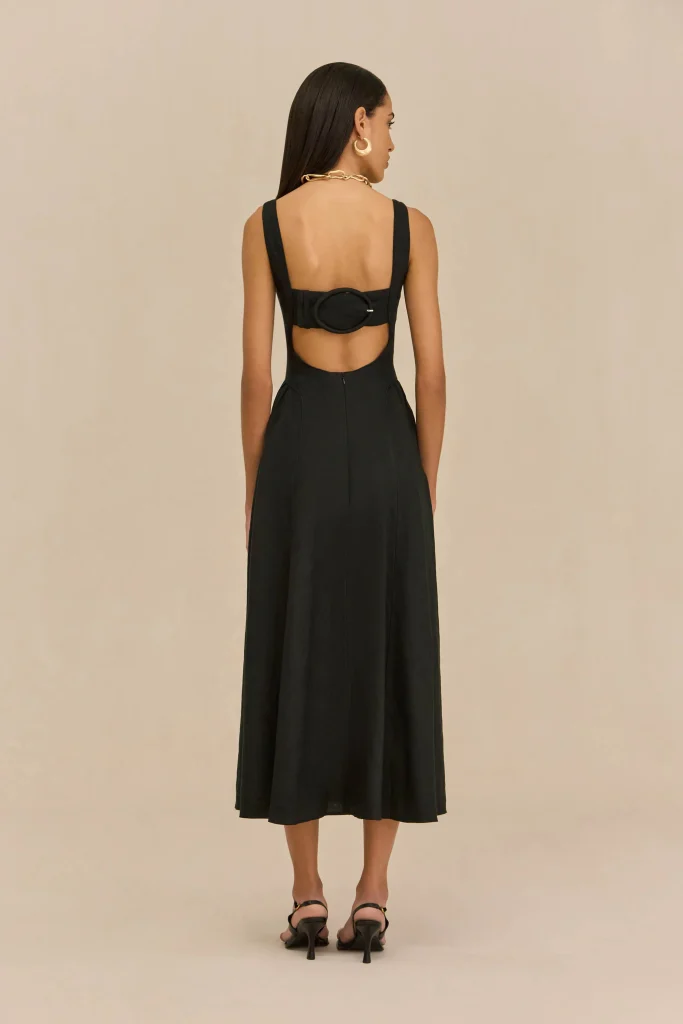 Back view of a woman in a black sleeveless dress with an open lower back and a large oval belt, wearing a chunky gold necklace and hoop earrings.