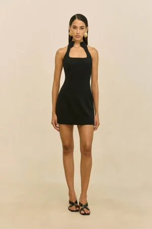 Full-body portrait of a woman in a black halter mini dress with large gold earrings, standing against a beige backdrop.