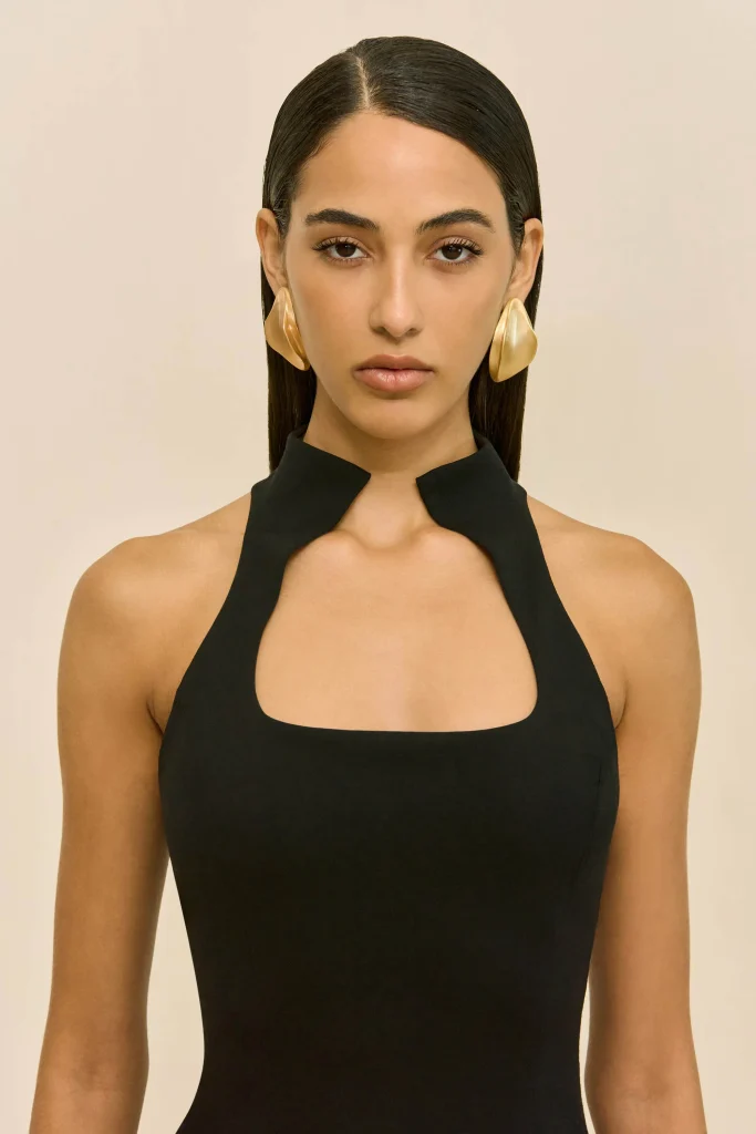 Woman facing camera in a black halter dress with large gold earrings against a neutral beige background.