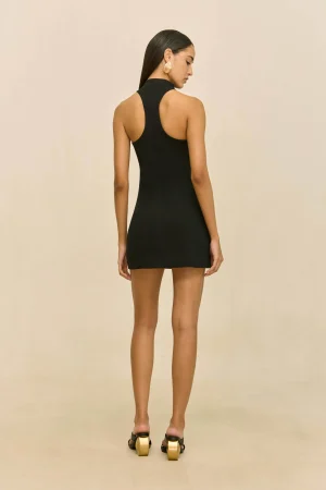 Back view of a woman wearing a black sleeveless mini dress with a high halter neckline and gold earrings, standing against a beige backdrop.