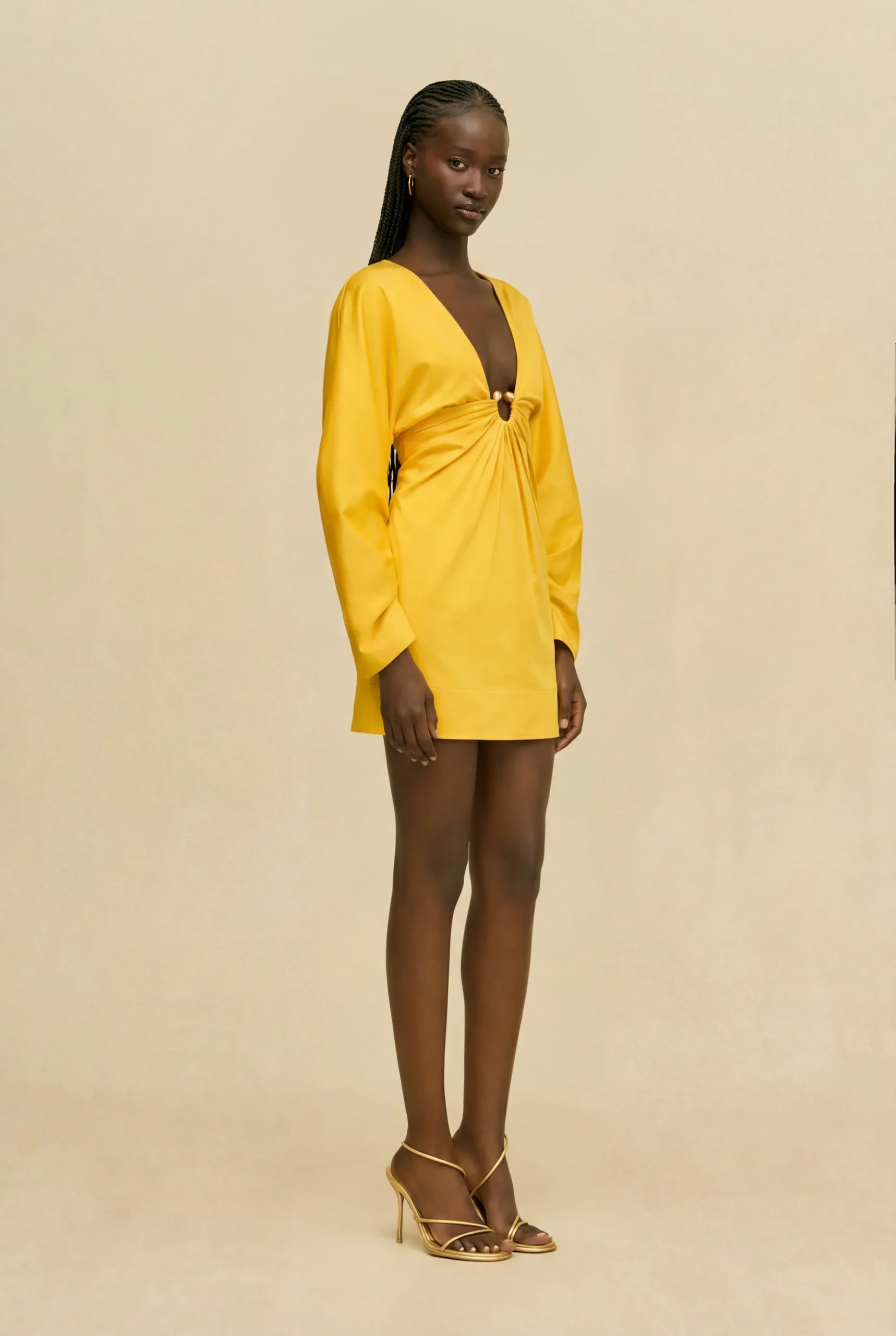 IMG_1522 - New Trend Model in a bright yellow mini dress with a deep V-neck and front knot, standing against a beige studio backdrop, wearing strappy heels at the ankles.
