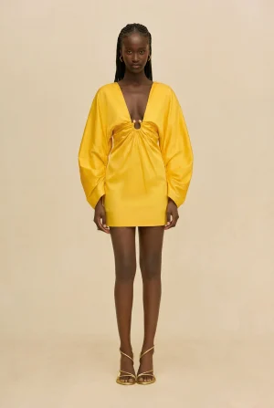 Full-body image of a woman in a bright yellow mini dress with a deep V neckline and gathered knot at the bust, wearing gold strappy sandals against a beige background.