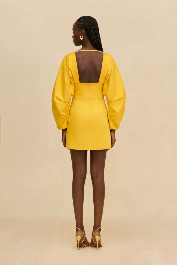 Woman standing with her back to the camera in a bright yellow dress with an open back and puff sleeves.