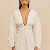IMG_1525 - New Trend Woman wearing a white, plunging-neckline dress with a ruched center knot and gold hoop earrings, standing against a beige background.