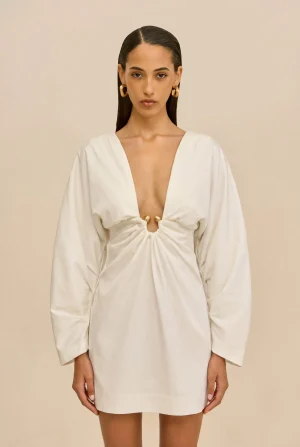 Woman wearing a white, plunging-neckline dress with a ruched center knot and gold hoop earrings, standing against a beige background.