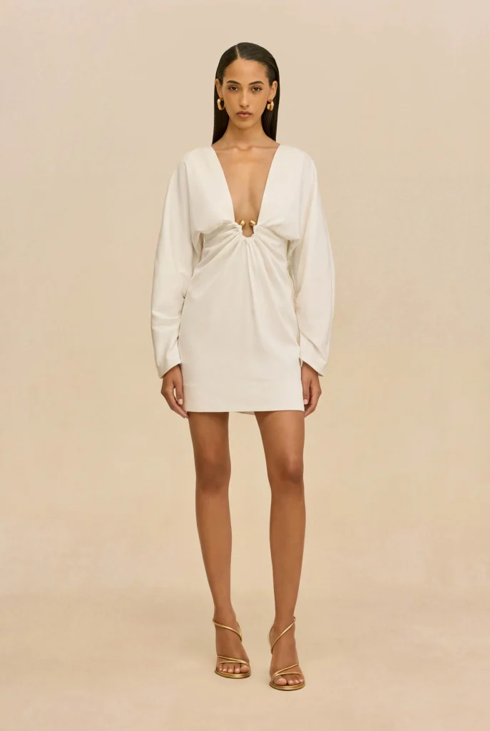 Model wearing a white deep-V mini dress with a front twist knot and long sleeves, gold hoop earrings, and strappy heels against a beige backdrop.