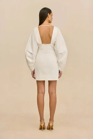 Back view of a woman in a white open-back dress with dramatic puff sleeves and gold strappy heels against a beige background.