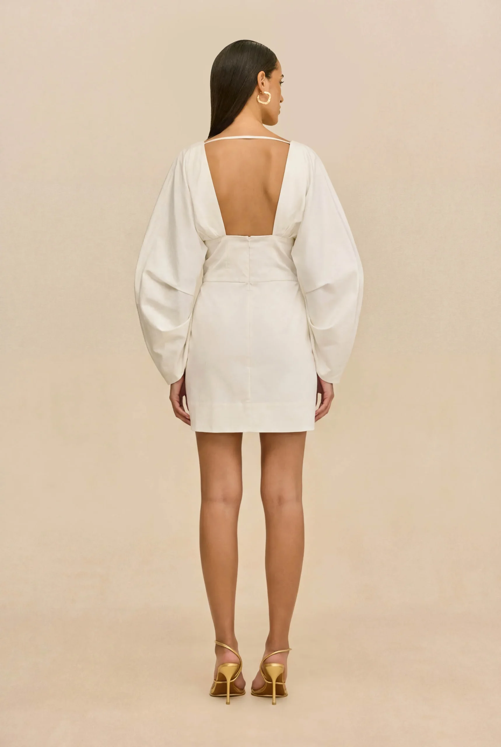 IMG_1528 - New Trend Back view of a woman in a white open-back dress with dramatic puff sleeves and gold strappy heels against a beige background.
