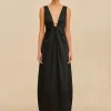 IMG_1529 - New Trend Woman standing in a black sleeveless dress with a deep V neckline and gold accents, against a beige backdrop, wearing open-toed sandals.