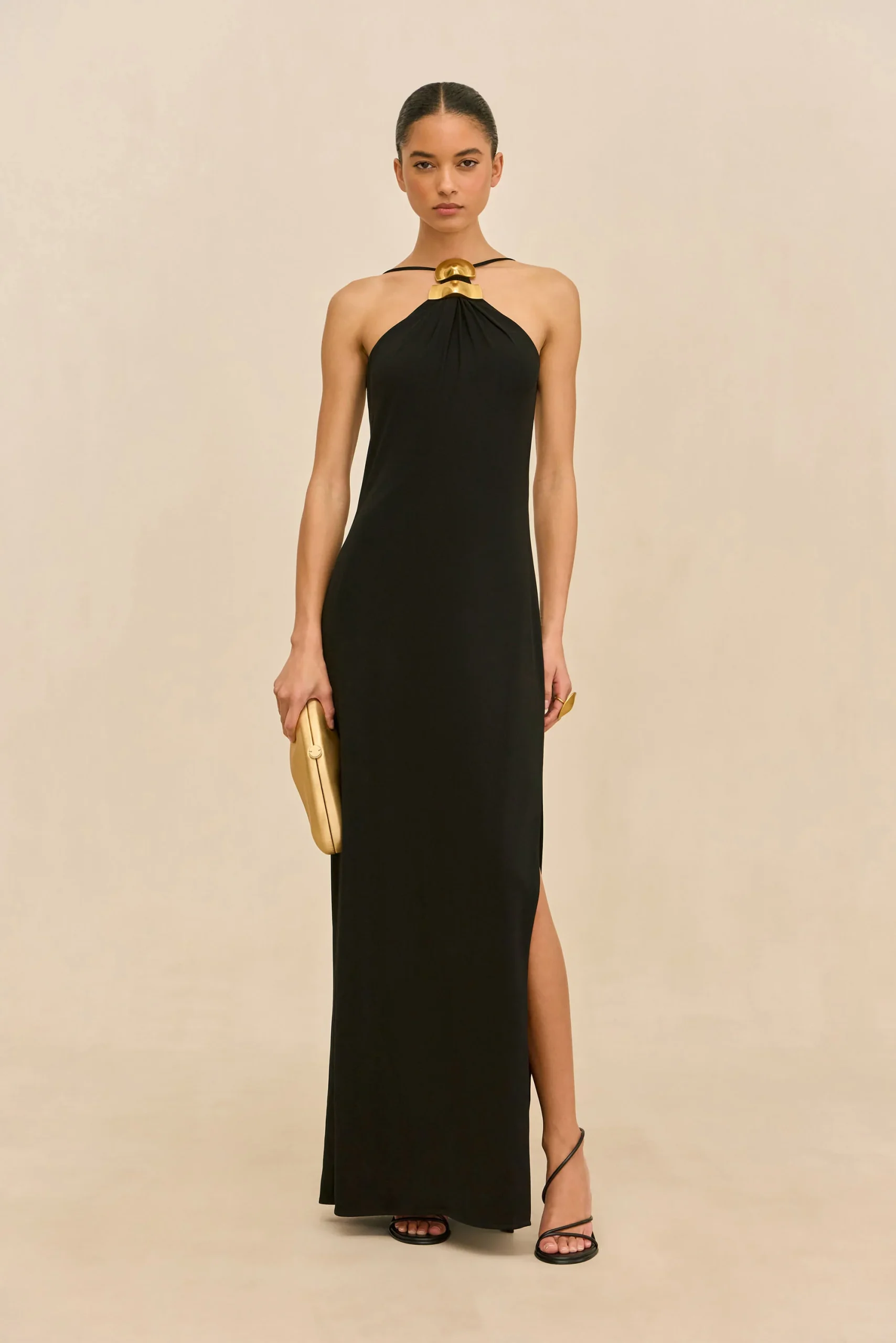 IMG_1533 - New Trend Model wearing a long black halter-neck gown with a gold neckpiece and matching gold clutch, standing against a beige background.