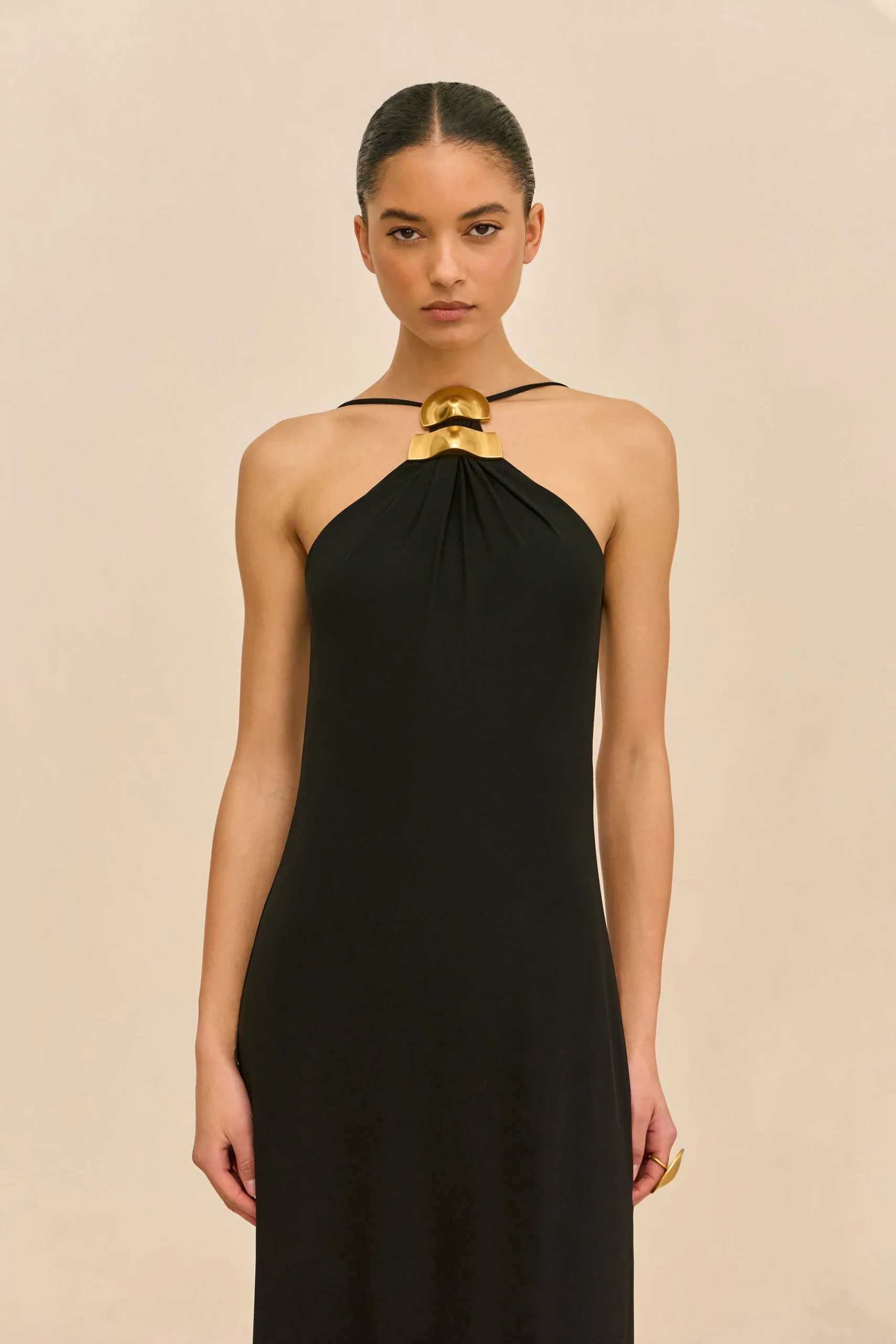 IMG_1534 - New Trend Model wearing a black halter dress with a bold gold geometric necklace, standing against a beige background.