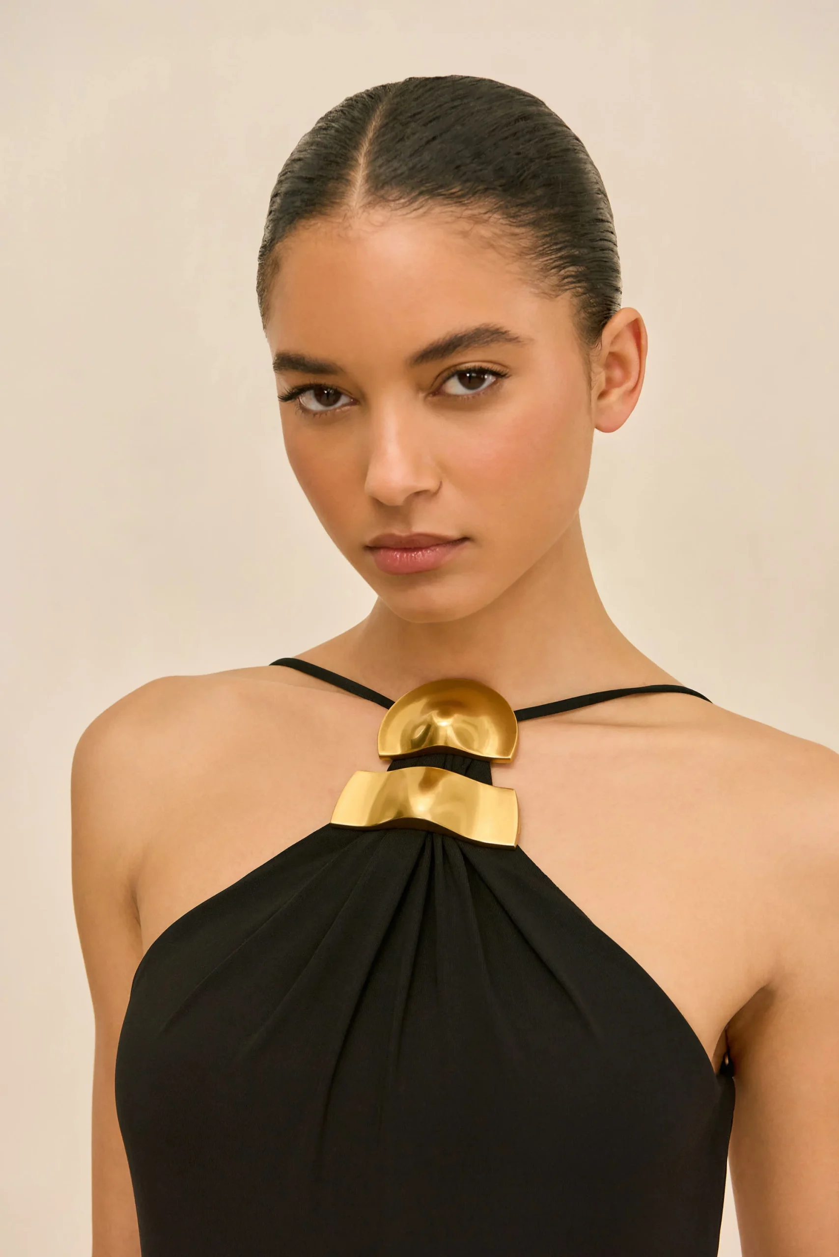 IMG_1535 - New Trend Portrait of a woman with slicked-back hair wearing a black halter dress and large gold statement necklace against a beige background, looking at the camera.
